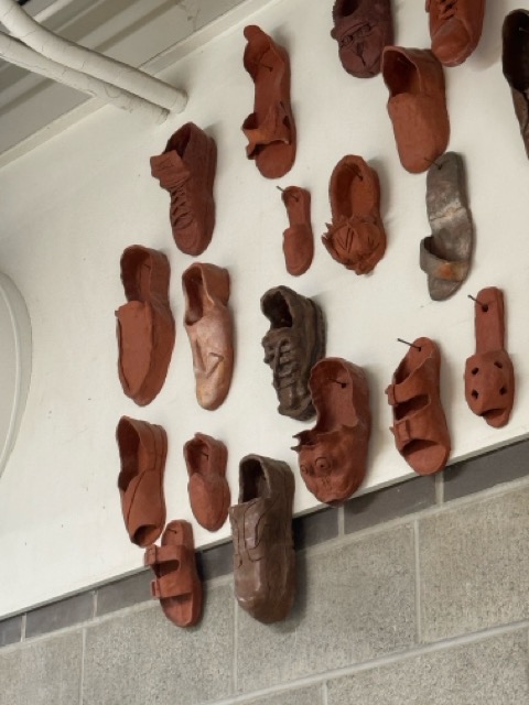 This image shows multiple shoes hung on the wall and arranged artistically, made my Sheridan ceramic students, they appear to be made of clay that was fired in the kiln to retain the shape however without glaze, making them appear to have a rougher texture.